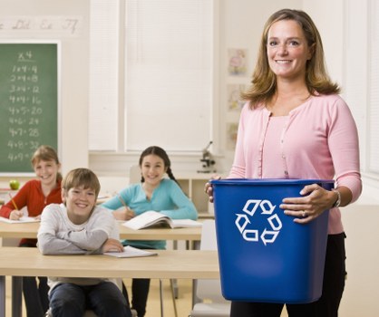 A woman in a pink blouse holding a blue plastic rubbish bin with a recycling symbol printed on it standing at the front of a classroom. In the background, three children are seated at desks, smiling and looking towards the camera. The classroom has white walls, a chalkboard with mathematical equations on the left side, and a window with closed blinds behind them. The setting suggests a school environment, and the presence of a rubbish bin relates to waste management and recycling activities, fitting with local rubbish removal services in Penge. The scene is well-lit, with natural light coming through the window, and the focus is on the woman and the bin she is holding, with the children and classroom environment providing contextual details indicative of an educational setting in the area.