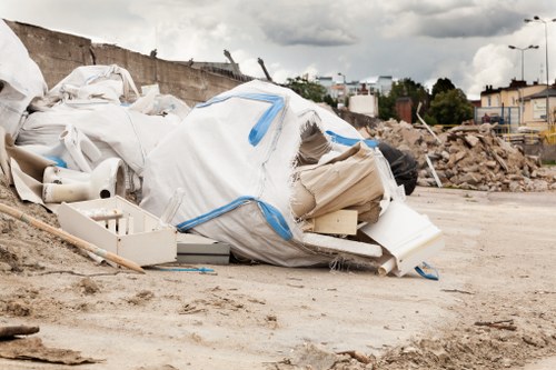 A large pile of white and beige waste materials, including plastic pipes, cardboard, and foam, is gathered on a construction site or cleared outdoor area. The debris appears to be construction or renovation refuse, with some items loosely stacked and partially covered by a white plastic bag or tarp reinforced with blue straps. In the background, there is a partially demolished wall with exposed bricks and rubble, along with a glimpse of buildings and trees further in the distance. The ground is uneven and dusty, typical of a site undergoing building or clearance work. The overall scene suggests waste removal activity in progress, consistent with services such as those provided by Penge House Clearance, which operates in the Penge area, within the postal code region of South London.