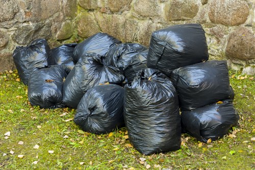 A collection of multiple black plastic rubbish bags filled with waste, resting on a patch of grass with scattered fallen leaves, positioned against a rustic stone wall. The bags are slightly shiny and appear to be securely tied at the tops, with some bags leaning on others, forming a loose cluster. The environment suggests an outdoor area, possibly part of a property or garden in Penge, with natural light illuminating the scene. The background consists of uneven stones with moss growth, indicating an exterior setting typical for rubbish collection and clearance services offered by Penge House Clearance. The image captures the typical appearance of waste ready for removal, emphasizing the need for professional rubbish disposal in the local area.