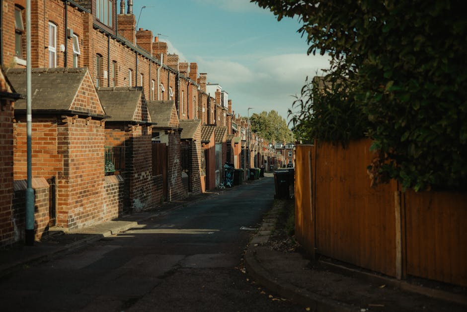 A narrow alleyway between two buildings with brick walls on the left and corrugated metal fencing on the right, both covered in graffiti and spray paint. The ground is uneven, muddy, and littered with