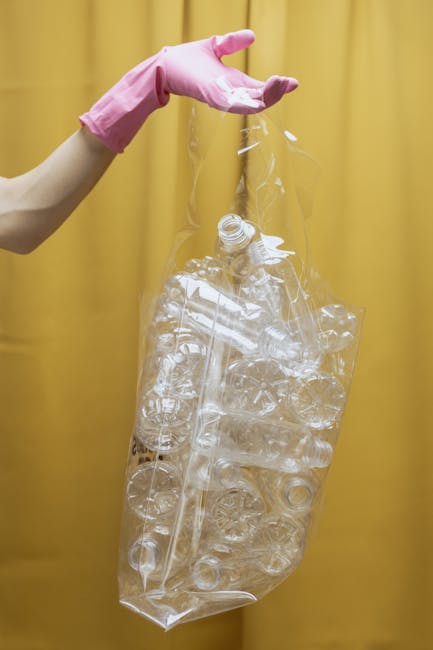 A person wearing a pink rubber glove is holding a large, clear plastic bag filled with numerous empty transparent plastic bottles, some crushed and others intact. The bags and bottles are set against 