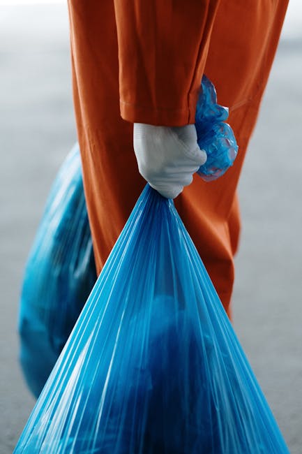 A person wearing a pink rubber glove is holding a large, clear plastic bag filled with numerous empty transparent plastic bottles, some crushed and others intact. The bags and bottles are set against 